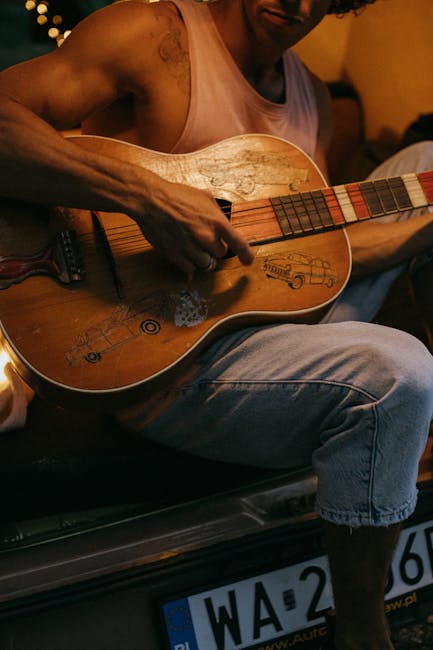A young man plays a guitar outdoors, sitting by a camper van, enjoying a musical moment.
